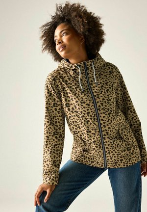 Donna con capelli ricci che indossa una felpa beige e nera con zip e stampa leopardata e jeans blu, in piedi con una gamba piegata e guarda di lato.