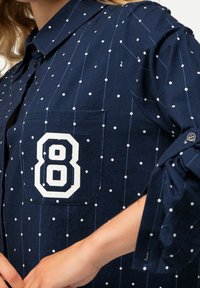 Navy blue shirt with white polka dots, featuring a chest pocket and a large white "8" patch. Buttoned collar with rolled sleeves.