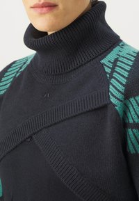 Temno modra turtleneck pulover s rebrastim ovratnikom, ki ima teal geometrijske vzorce na ramenih, izdelan iz mehkega, teksturiranega pletenega materiala.