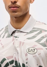 Κοντινό πλάνο ενός ατόμου που φορά μια ανοιχτό γκρι πουκάμισο με γιακά, διακοσμημένο με αφηρημένα φυτικά μοτίβα και ένα οβάλ σήμα λογότυπο EA7 Emporio Armani.