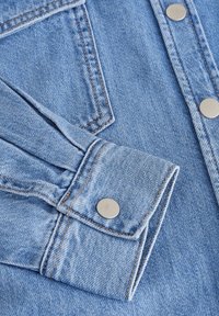 Tessuto di denim azzurro chiaro con cuciture visibili e bottoni a pressione in metallo argentato. L'immagine si concentra su un dettaglio del risvolto piegato.