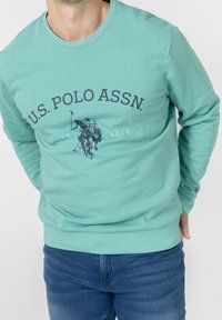 Man wearing a teal U.S. Polo Assn. sweatshirt with a horse and rider logo, paired with blue jeans on a white background.