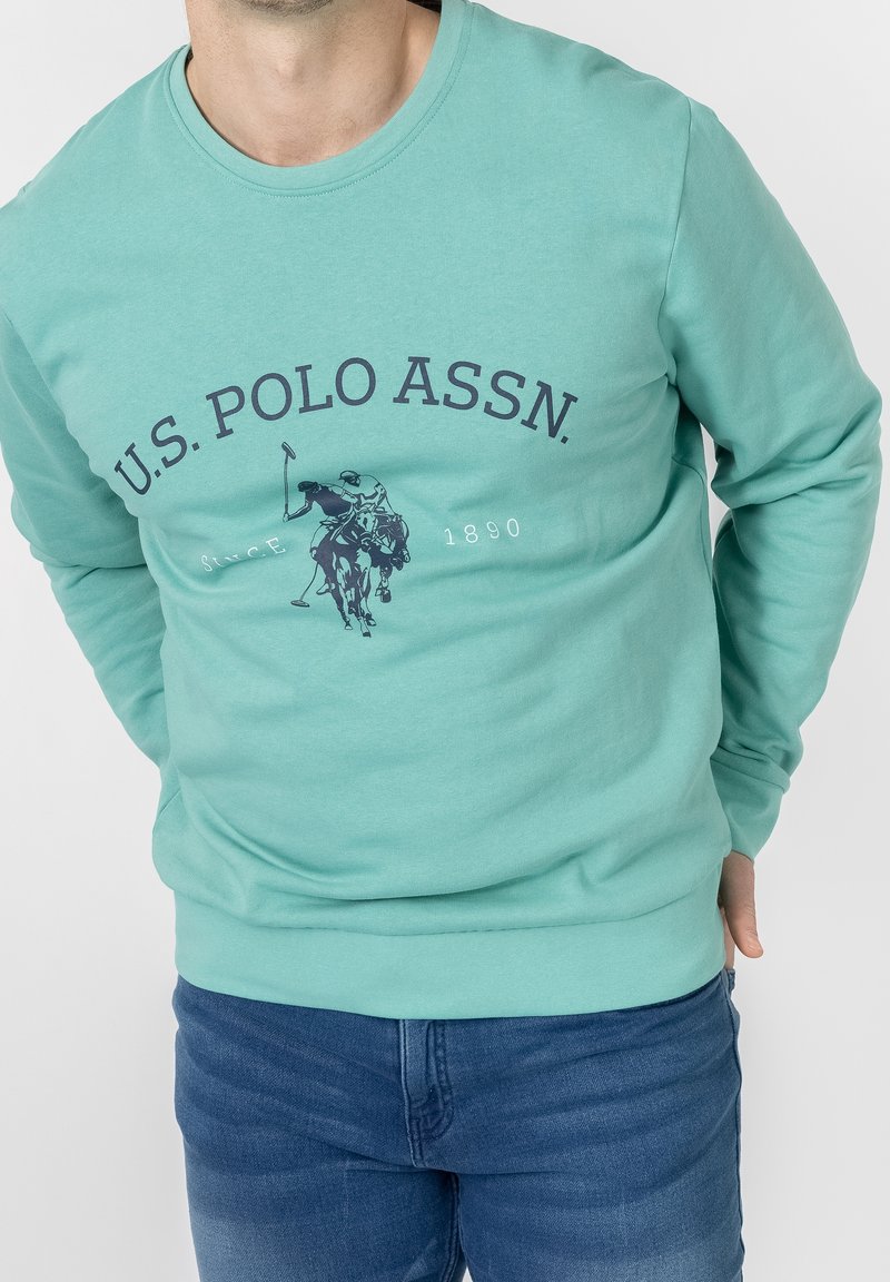 Man wearing a teal U.S. Polo Assn. sweatshirt with a horse and rider logo, paired with blue jeans on a white background.