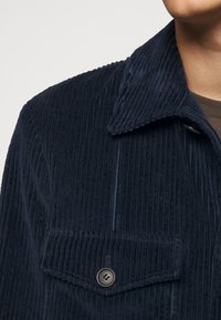 Zbliżenie na kieszeń i kołnierz granatowej katanowej kurtki noszonej na brązowej koszuli, ukazujące guzik i pionowe prążki materiału.