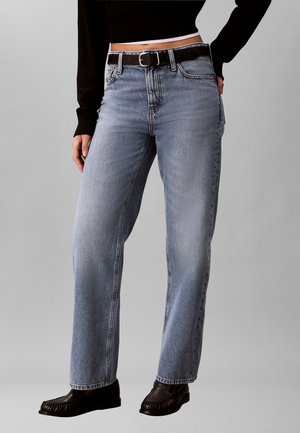 Personen har lyseblå jeans med lige ben og sort bælte på, sort langærmet crop top og sorte sko mod en ensfarvet baggrund.