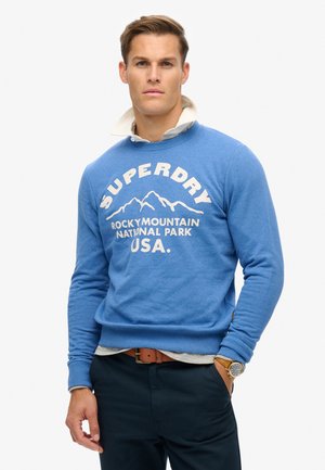 Sudadera azul de cuello redondo con puños y dobladillo acanalados, que presenta un gráfico de montaña blanca y el texto "SUPERDRY ROCKY MOUNTAIN NATIONAL PARK USA".