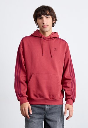 Rote Kapuze mit einer Fronttasche, strukturiertem Material und drei lila Streifen an jedem Ärmel. Mit gesticktem Logo auf der Brust.