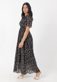 Maxi-jurk in zwarte bloemenprint met korte mouwen, elastische taille en lichte stof. Gecombineerd met witte haksandalen.