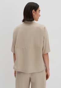 Camicia beige a maniche corte con vestibilità rilassata, texture liscia e dettaglio della cucitura orizzontale sul retro. Abbinata a pantaloni coordinati.