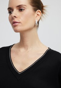 Pull à col en V noir avec une bordure argentée, présentant une texture lisse. Modèle portant une boucle d'oreille argentée, mettant en avant un design texturé.