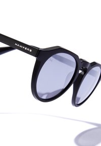 Črna sončna očala z ostrimi okvirji iz mat plastike, z temnimi lečami in kovinskim poudarkom z imenom blagovne znamke "HAWKERS".