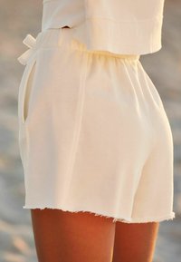 Lys beige elastisk talje shorts med sidelommer og rå kant, båret af en person, der står udendørs på sand.