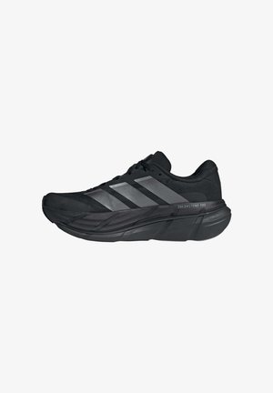 Zapatilla de correr Adidas negra con suela gruesa y tres rayas reflectantes, etiquetada como Dreamstrike Pro en la entresuela.