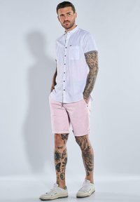 White short-sleeve button-up shirt with a front pocket, paired with light pink shorts. White sneakers complete the outfit, showcasing tattooed legs.