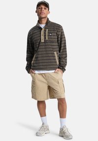 Πουλόβερ σκουρό καφέ με οριζόντιες ρίγες, ελαφρύ, με ψηλό γιακά, τρία κουμπιά και τσέπη στο στήθος· συνδυασμένο με χακί cargo σορτς.