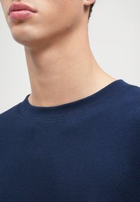 Námornícky modrý textúrovaný sveter s rebrovaným výstrihom, zobrazujúci detailný pohľad na golier a plece. Mäkký materiál.