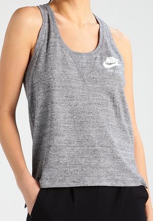 Kobieta w szarej, bezrękawnikowej bluzce Nike z logo „Just Do It”, z rękami w kieszeniach czarnych spodni, na jednolitym tle.