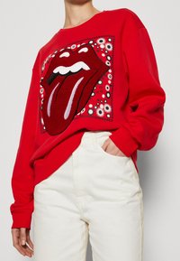 Rotes Sweatshirt mit einem großen, bestickten Rolling-Stones-Logo und einem markanten Zunge-Design, umgeben von einem floralen Muster. Kombiniert mit weißen Jeans.