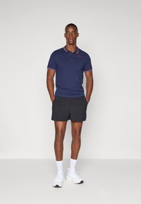 Marineblå poloskjorte med røde og hvite krageakenter, kombinert med svarte shorts. Modell som bruker hvite joggesko og ankelstrømper.
