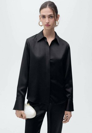 Black satin long-sleeve shirt with a pointed collar and curved hem, paired with a white oval clutch featuring a gold clasp.