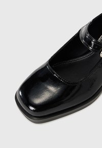 Chaussure Mary Jane en cuir verni noir avec un bout carré, une bride de cheville et une légère texture sur la surface, dotée d'une semelle plate.