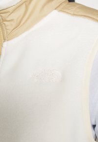Bílé fleecové vesty s béžovým ramenním panelem. Na přední straně je výšivka loga v bílé barvě. Má hladkou texturu a lehký design.
