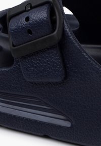 Bride de sandale texturée bleu foncé avec boucle noire réglable et détail de semelle rainurée sur une chaussure à bout fermé.