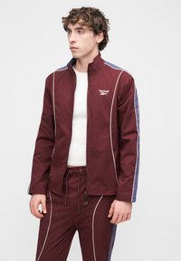 Bordó cipzáras dzseki fehér csíkozással és tengerészkék oldalsó panelekkel. Könnyű anyagból készült, magas gallérral és karcsúsított szabással.