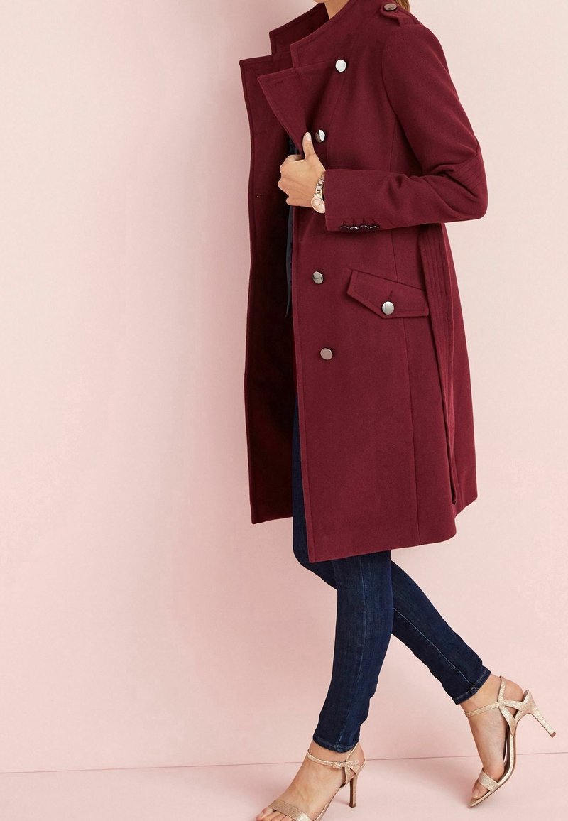 Femme portant un manteau croisé bordeaux, un jean skinny foncé, une montre et des sandales à talons beige, posant devant un fond rose pâle.