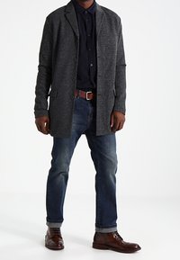 Grå strik lang blazer med reverskrave, sort button-up skjorte, blå jeans og brune lædersko. Glat tekstur og skræddersyet pasform.