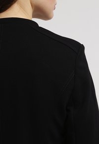 Černá bunda s hladkým povrchem, s minimalistickým designem, viditelnými ramenními švy a jemnými detaily šití.