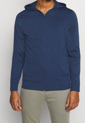 Mann trägt eine dunkelblaue Kapuzenjacke mit Reißverschluss und hellgraue Hose, steht vor einem einfarbigen hellen Hintergrund.