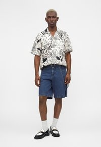 Homme portant une chemise à manches courtes à motif noir et blanc, un short en jean avec une chaîne, des chaussettes blanches et des mocassins noirs, debout devant un fond blanc.