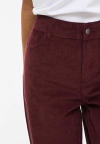 Close-up van een persoon die een hooggetailleerde bordeauxrode ribfluwelen broek en een ingestopt wit shirt draagt, met de focus op de taille en het bovenbeen.