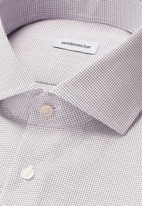 Primo piano di una camicia bianca con un fine motivo a griglia nera, colletto appuntito, due bottoni bianchi e un'etichetta del marchio visibile all'interno del colletto.
