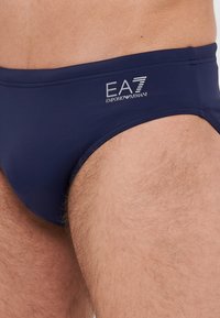 Fatos de banho em azul marinho, feitos de material elástico, com um logotipo branco da EA7 Emporio Armani na cintura, textura suave e design ajustado.