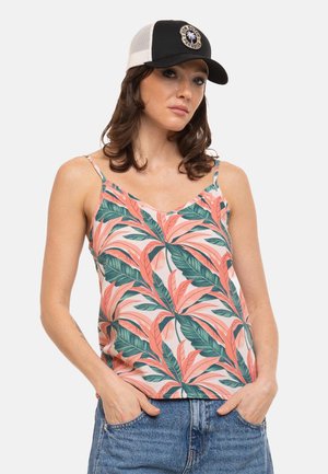 Femme portant une casquette noir et blanc, un top sans manches avec un motif de feuilles roses et vertes, et un jean bleu avec les mains dans les poches.