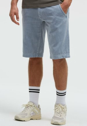 Mann trägt hellblaue knielange Shorts, weiße Socken mit schwarzen Streifen und beige Sneaker, steht mit einer Hand in der Tasche.