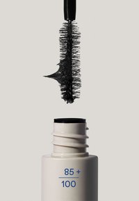 Ett mascaraborste täckt med svart mascara hålls ovanför en öppen vit tub märkt "85 + 100" mot en enkel bakgrund.