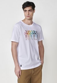 T-shirt in cotone bianco con un design grafico colorato di una bicicletta sul davanti. Maniche corte, collo tondo e vestibilità morbida.