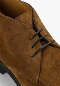 Brune ruskind ankelstøvle med en tekstureret finish, udstyret med sorte snørebånd, en rund tå og en subtil stakhælsdesign.