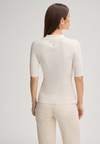 Donna con i capelli castani lisci, che indossa una maglietta aderente a maniche corte di colore crema a costine e pantaloni beige chiaro, vista da dietro.
