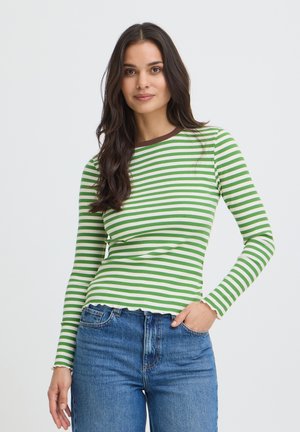 Femme aux longs cheveux foncés portant une chemise à manches longues rayée verte et blanche et un jean bleu, debout avec une main dans la poche sur un fond uni.