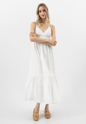 Frau mit langen blonden Haaren trägt ein ärmelloses weißes Maxikleid und gewebte beige Ballerinas, steht mit verschränkten Armen vor einem weißen Hintergrund.