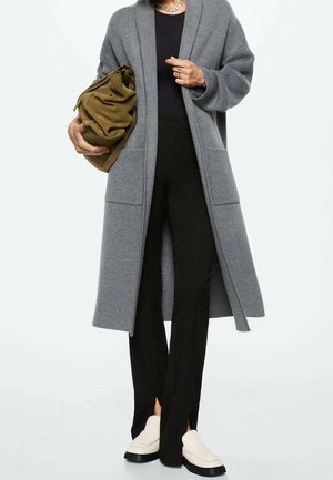 Osoba nosi długi szary płaszcz, czarne spodnie z przednimi rozcięciami, białe wsuwane buty, trzyma dużą brązową miękką torbę oraz nosi masywny naszyjnik z łańcucha.