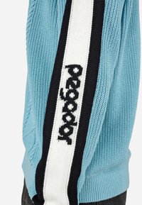 Šviesiai mėlynas megztinis su tekstūruotu raštu. Juodai baltas juostuotas akcentas su tekstu eina per rankovę. Džersio rankogaliai ties riešais.