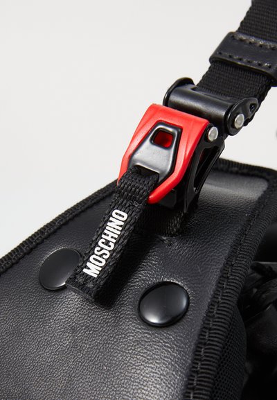 Sort tekstureret materiale med en blank finish, der har en rød plastikspænde, sort rem og et hvidt "Moschino" mærke.