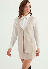 Beige väst med djup V-ringning, två framfickor, lager över en vit skjorta med knappar. Modellen bär en matchande kjol och knähöga stövlar.