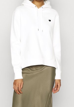 Persona con una sudadera blanca con cordones frontales y un pequeño emblema de corazón negro, combinada con una falda verde oliva.