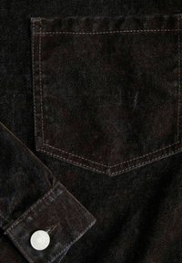 Close-up van donkere denim stof waarop een achterzak te zien is met contrasterende stiksels en een mouwomslag met een metalen knop.